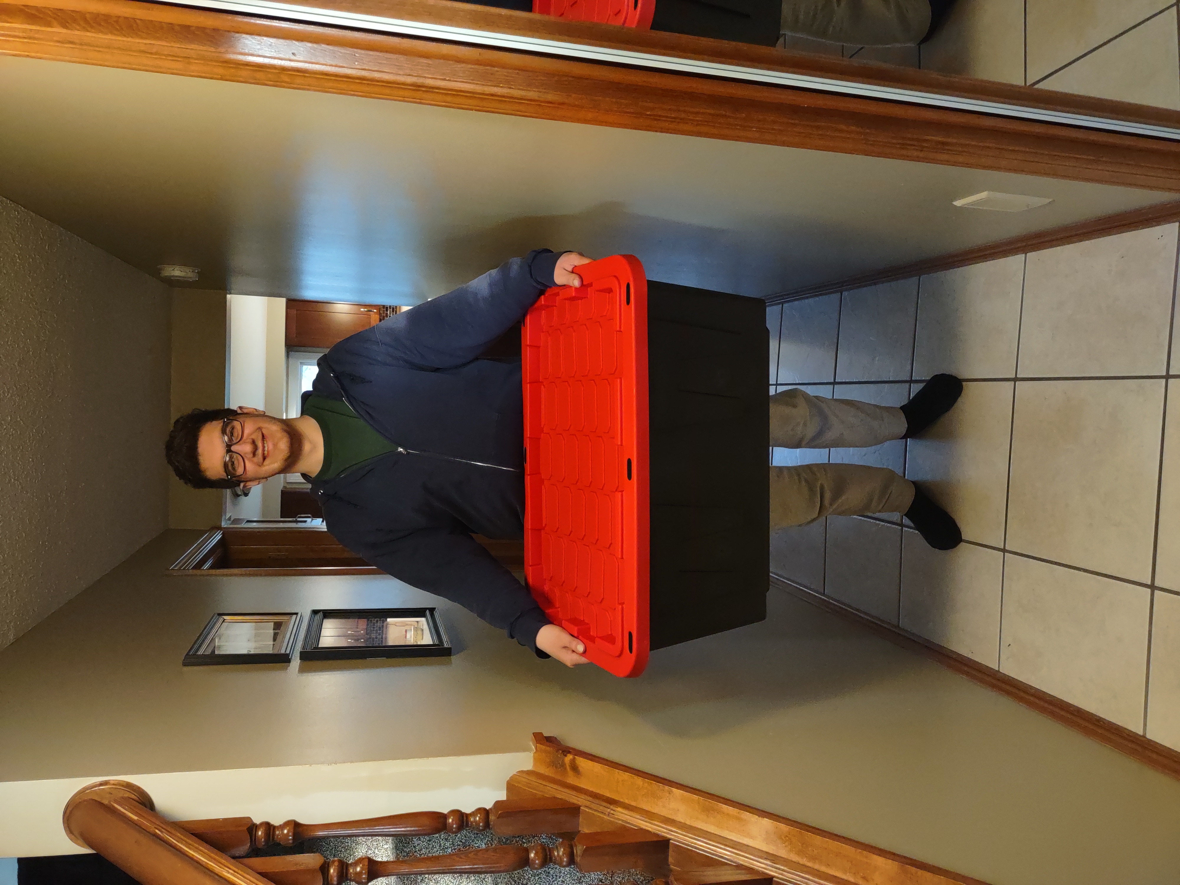 Smiling man holding a large black and red reusable plastic moving tote in a hallway